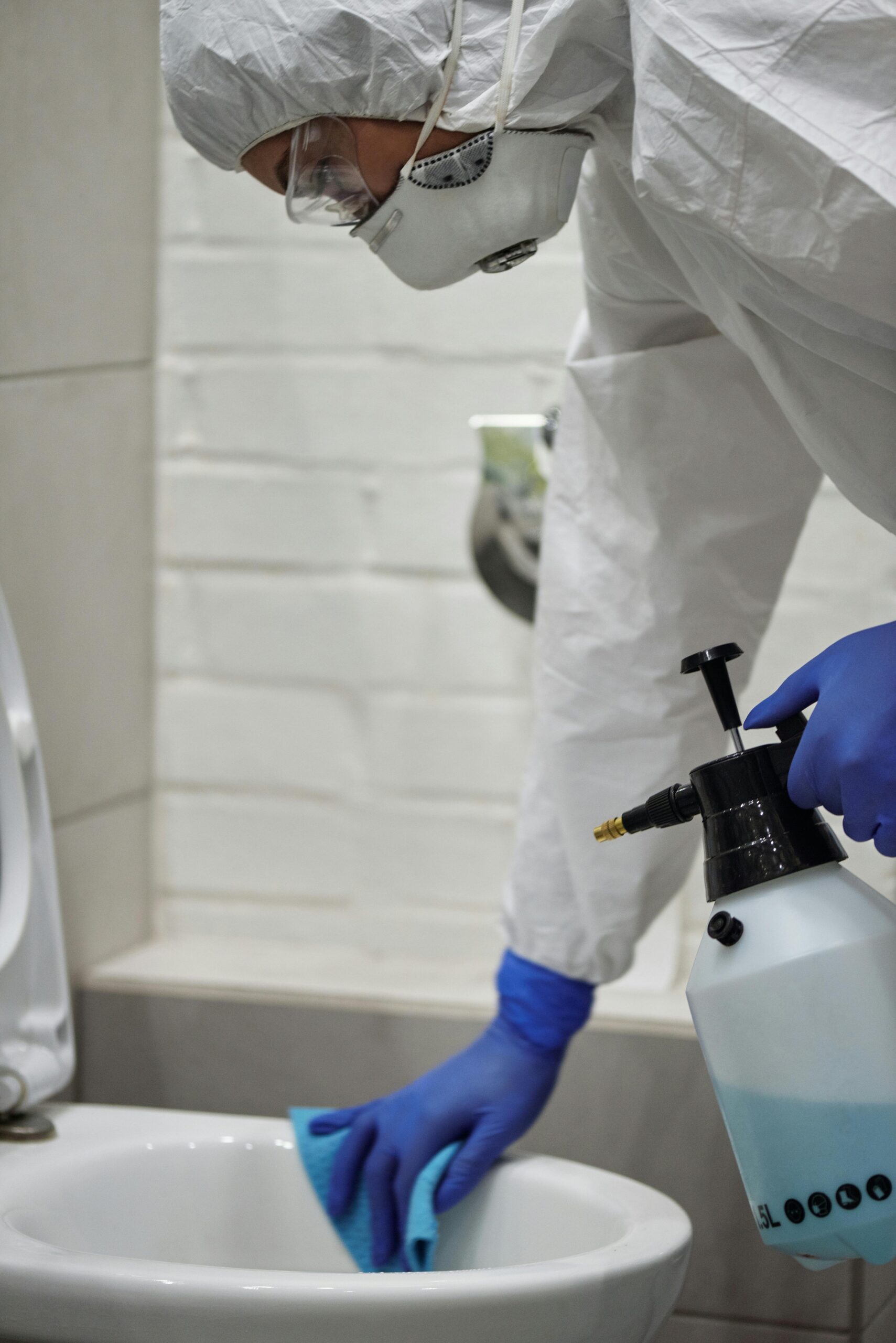 Person in protective gear cleaning and disinfecting a toilet in a bathroom environment.